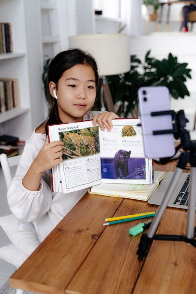 The Art of Drawing Readers In: Your attractive post title goes here Young girl displays book during online class using smartphone and tripod.
