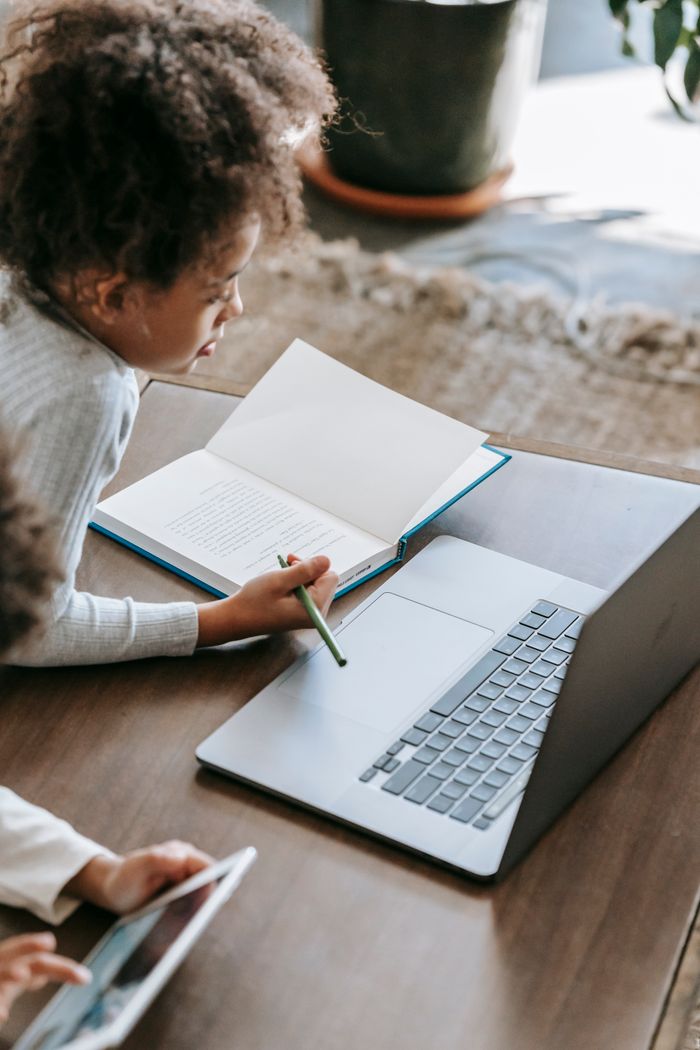 From above side view of concentrated African American girl with pen sitting at table with book and modern netbook while studying at home